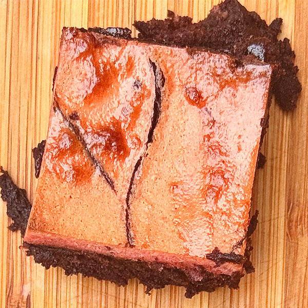 Chocolate brownie square with a glossy pink topping on a wooden board.