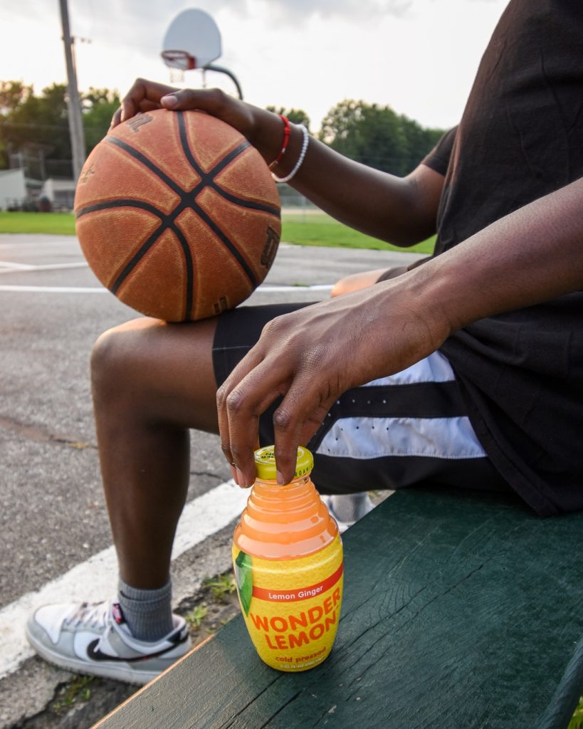 A person holding a basketball sits on a bench at an outdoor court with a bottle of Wonder Lemon juice beside them.