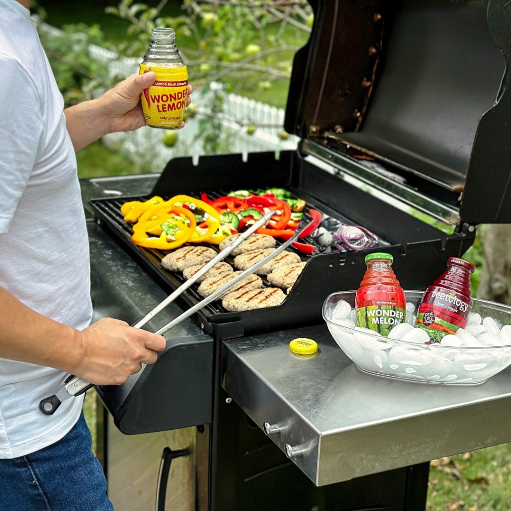 Man grilling veggies and chicken while holding Wonder Lemon drink, with more bottles chilling in ice beside the grill.