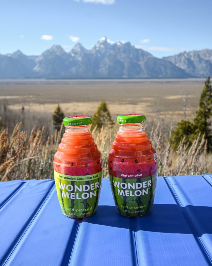 Two bottles of Wonder Melon sit on a blue table with a stunning mountain range and open landscape in the background.