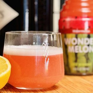 Fresh pink juice in a glass with lemon and a Wonder Melon bottle behind it.